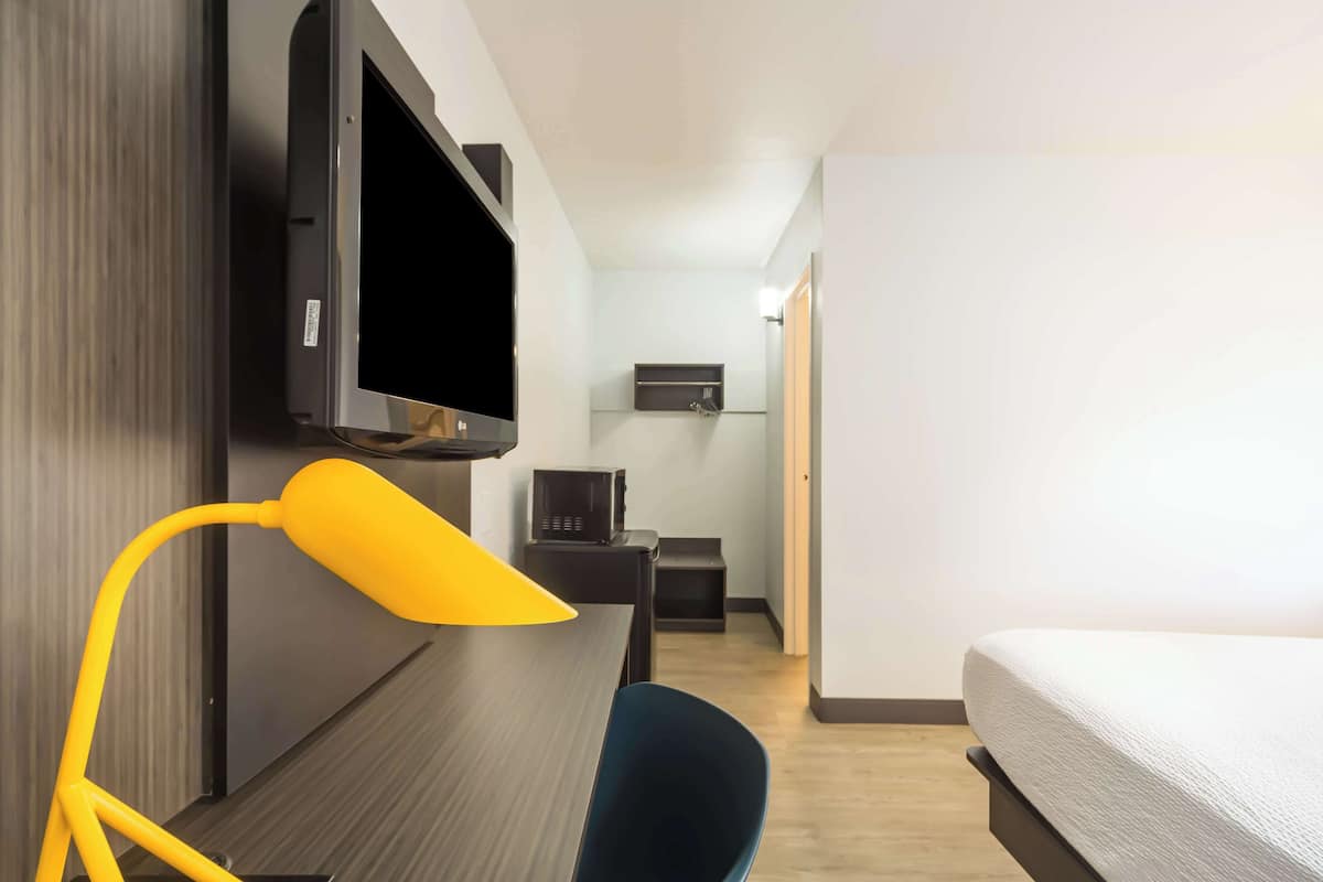 A hotel room with a bed, a desk with a chair, a TV mounted on the wall, and a microwave in the kitchenette area.