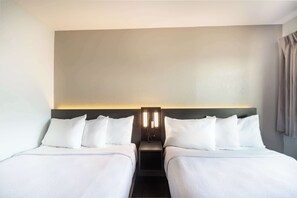 A hotel room with two beds, a nightstand with a lamp, and a plain wall.