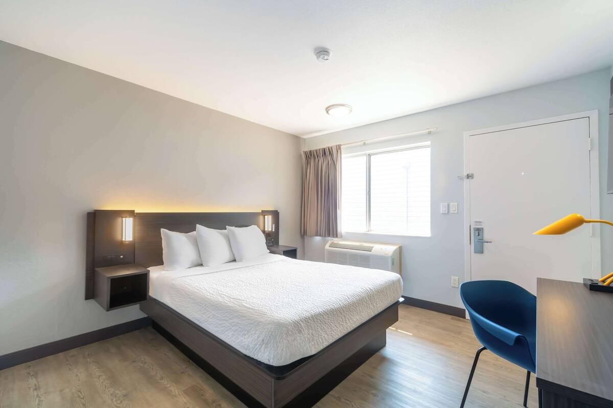 A hotel room with a large bed, bedside tables, a desk with a chair, a window with curtains, and a door.
