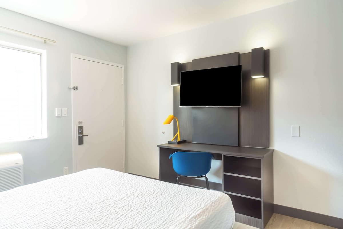 A bedroom with a bed, a desk with a chair, a TV mounted on the wall, and a door.