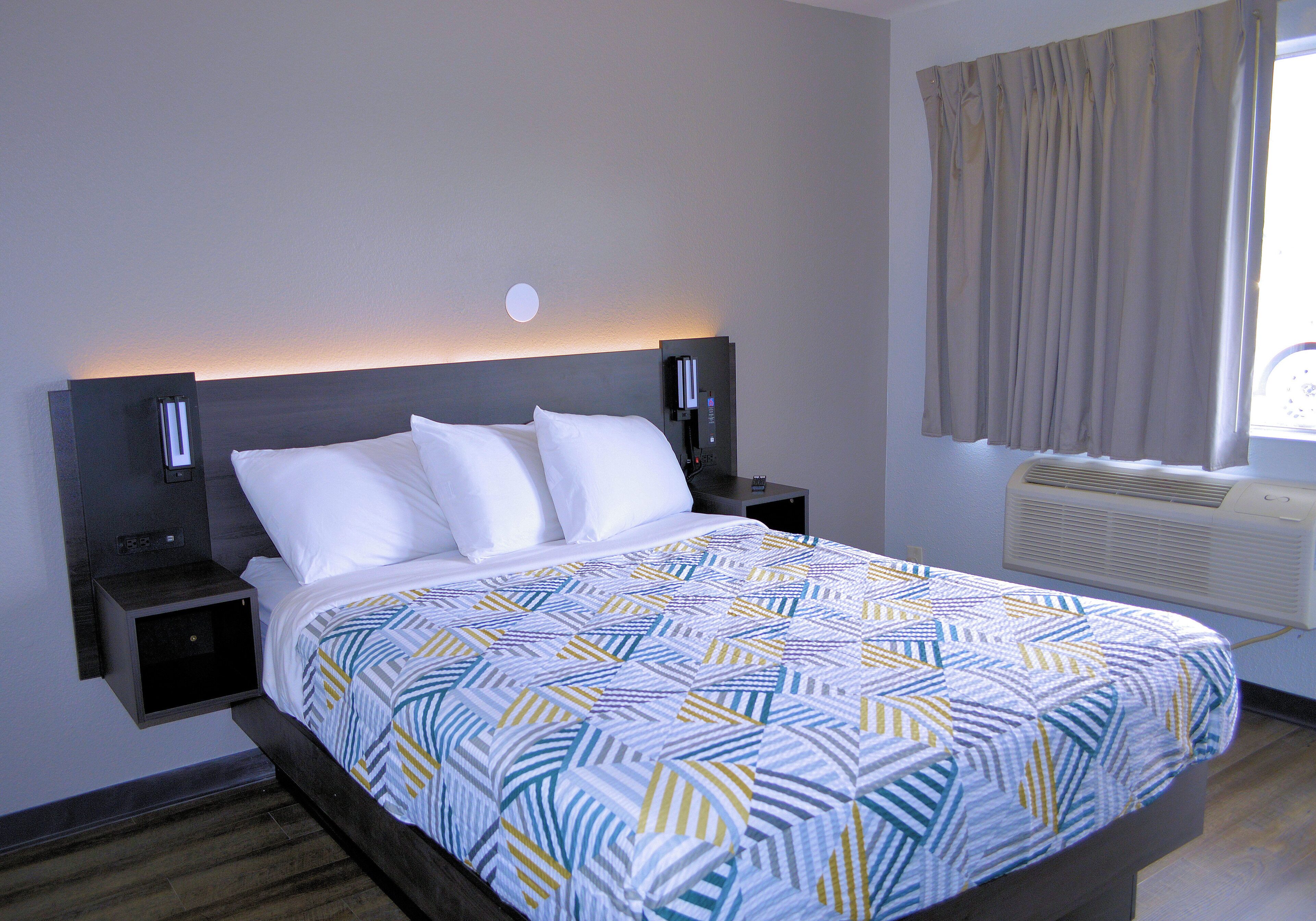 Een net opgemaakt bed met een beddengoed met patroon, twee nachtkastjes met lampen, een wandlamp en een raam met gordijnen.