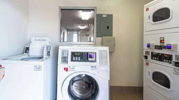 A laundry room with multiple washing machines and dryers, a control panel, and a door leading to another room.