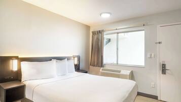 A hotel room with a large bed, bedside lamps, a window with curtains, an air conditioning unit, and a door.