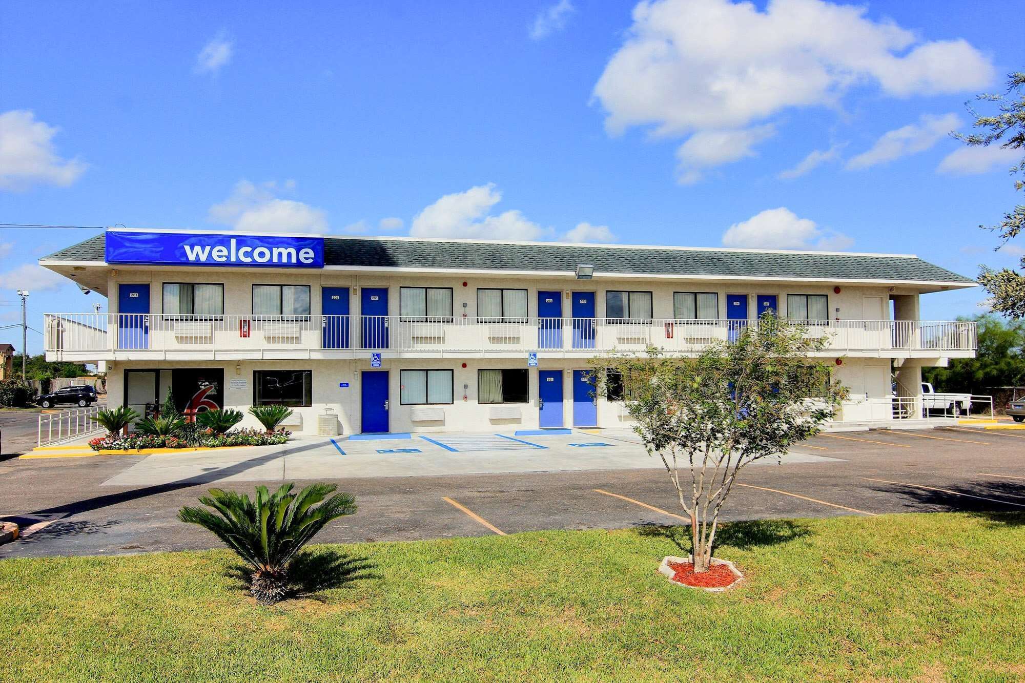 Un motel a due piani con un cartello 'Welcome', diverse porte blu e un parcheggio.