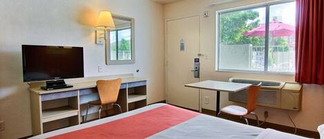 A hotel room with a bed, a desk with a TV, a chair, a window with curtains, and a heater.