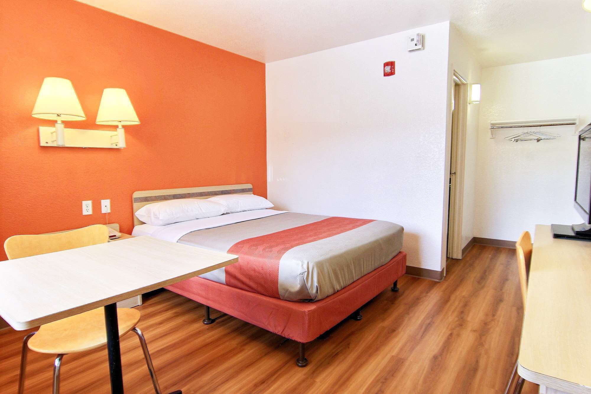A hotel room with a bed, a desk, a chair, and a table. There are two wall-mounted lamps on the left side of the bed.