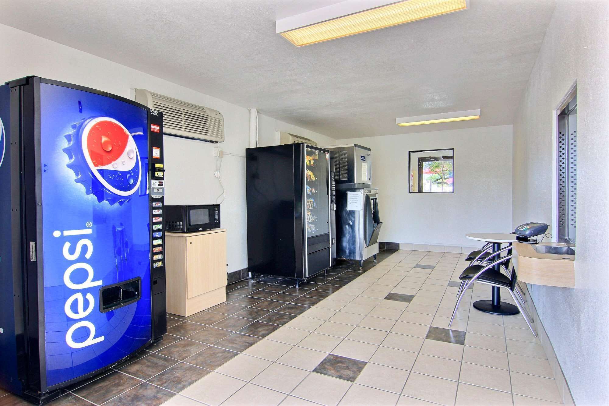 A hotel lobby with a Pepsi vending machine, a microwave, a refrigerator, and a small table with chairs.