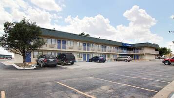 A motel with a parking lot, multiple cars parked, and a tree in the foreground.