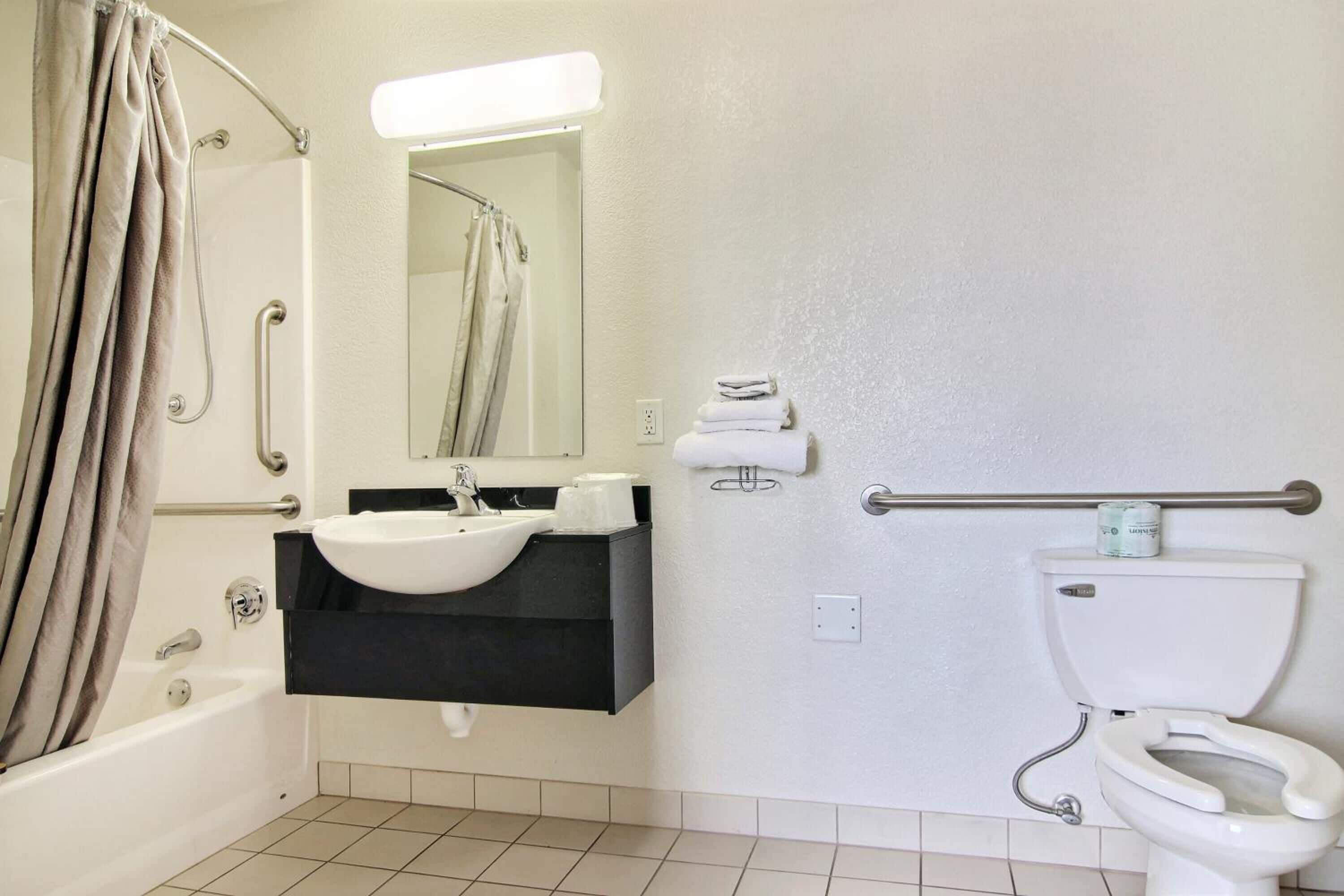 A bathroom with a bathtub, sink, mirror, toilet, and handrails.