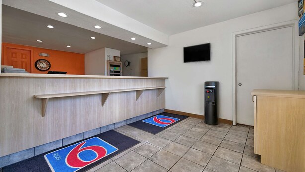 A hotel reception area with a wooden counter, a clock, a trash bin, and a door. - Motel 6 Alamogordo, NM (Alamogordo)