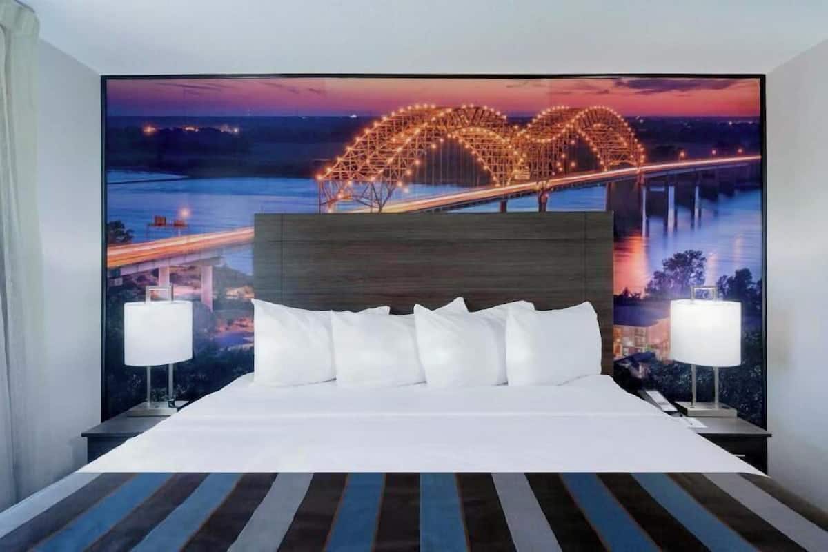 A hotel room with a large bed, two bedside lamps, and a mural of a bridge at dusk.