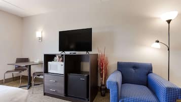 A hotel room with a bed, a desk with a chair, a black minibar, a television on a stand, a blue patterned sofa, and a floor lamp.