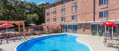 A swimming pool with a sunshade, lounge chairs, and a brick building in the background.