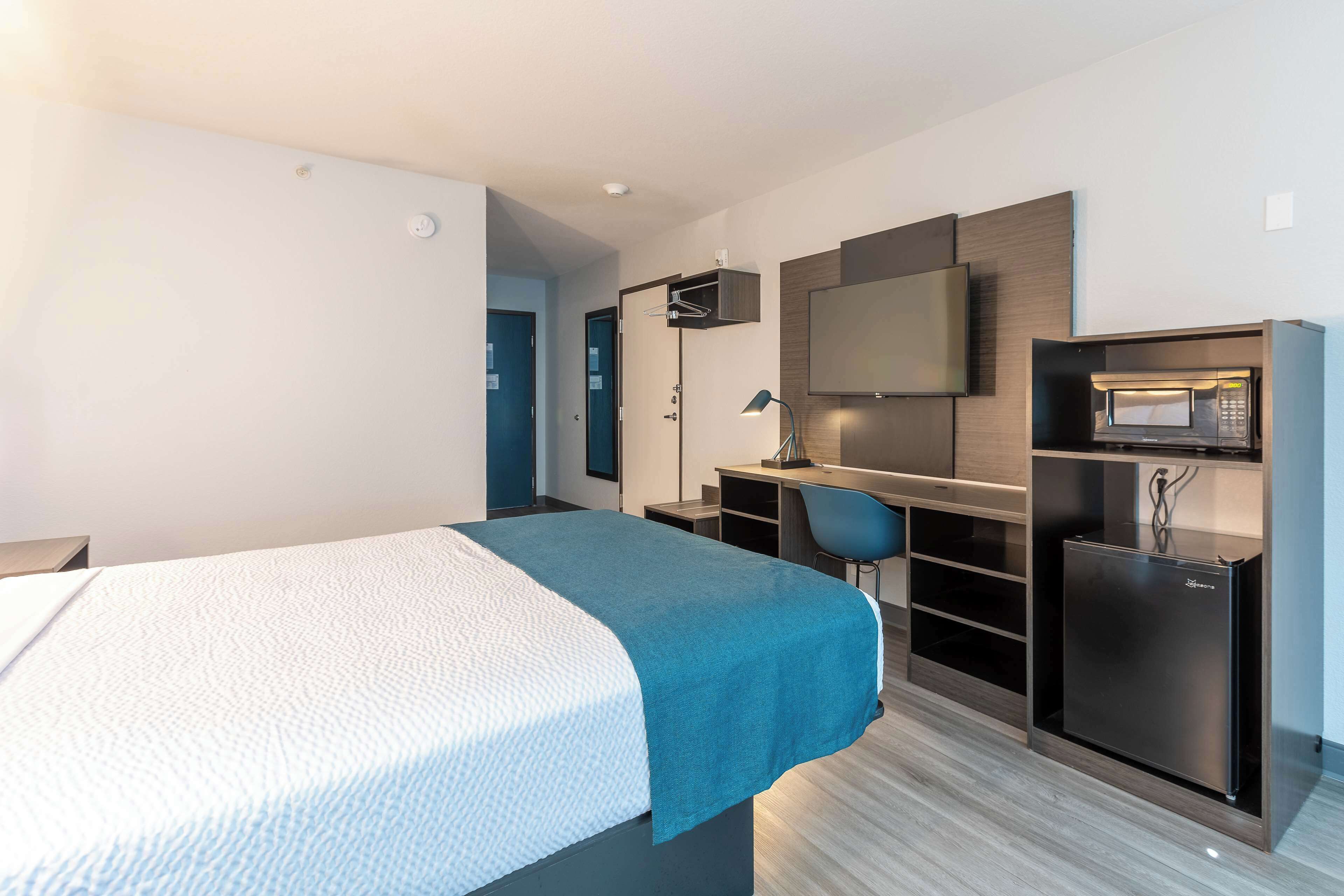 Quarto de hotel moderno com cama, escrivaninha, cadeira, TV e micro-ondas embutido.