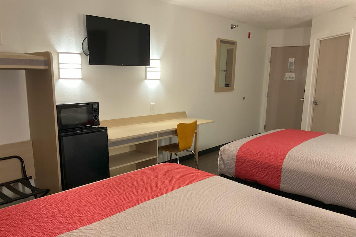 A hotel room with two beds, a desk, a chair, a TV, and a refrigerator.