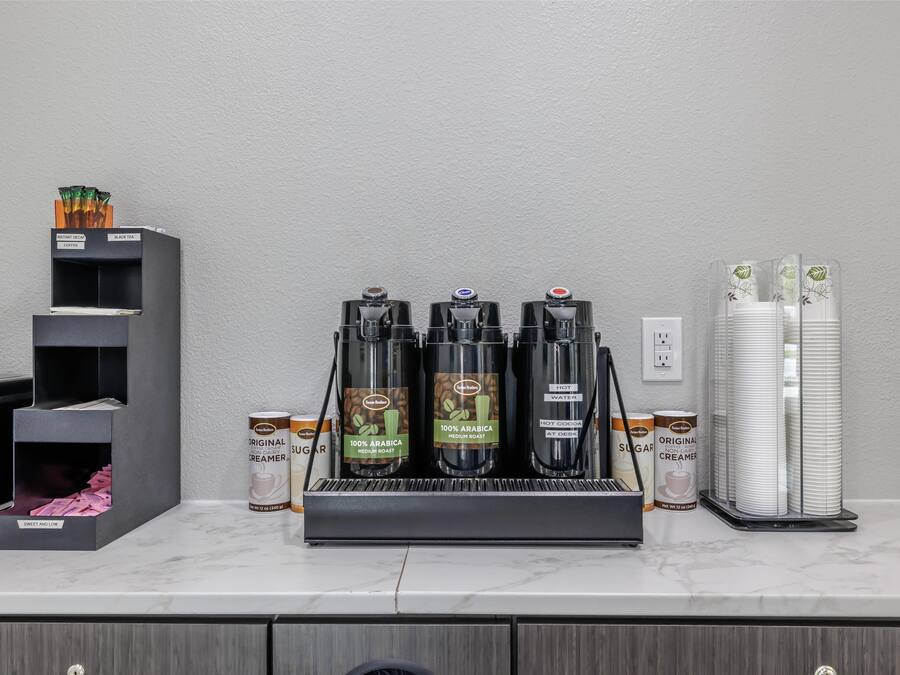 A coffee station with three coffee dispensers, sugar and creamer dispensers, and a stack of white cups on a marble countertop.