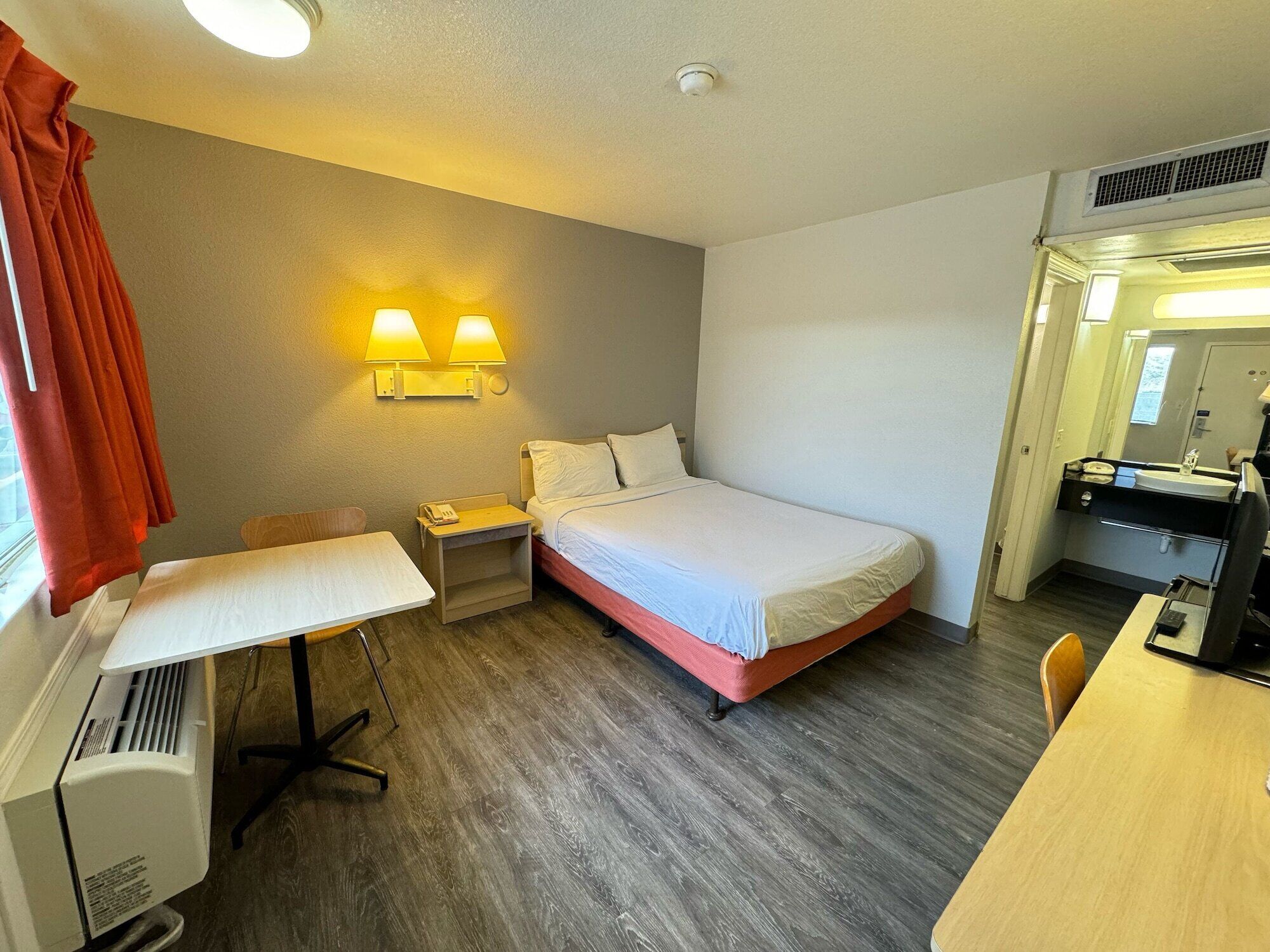 Ein Hotelzimmer mit Bett, Schreibtisch, Stuhl, einem kleinen Tisch, einer Heizung und einem Badezimmer mit Waschbecken und Spiegel.