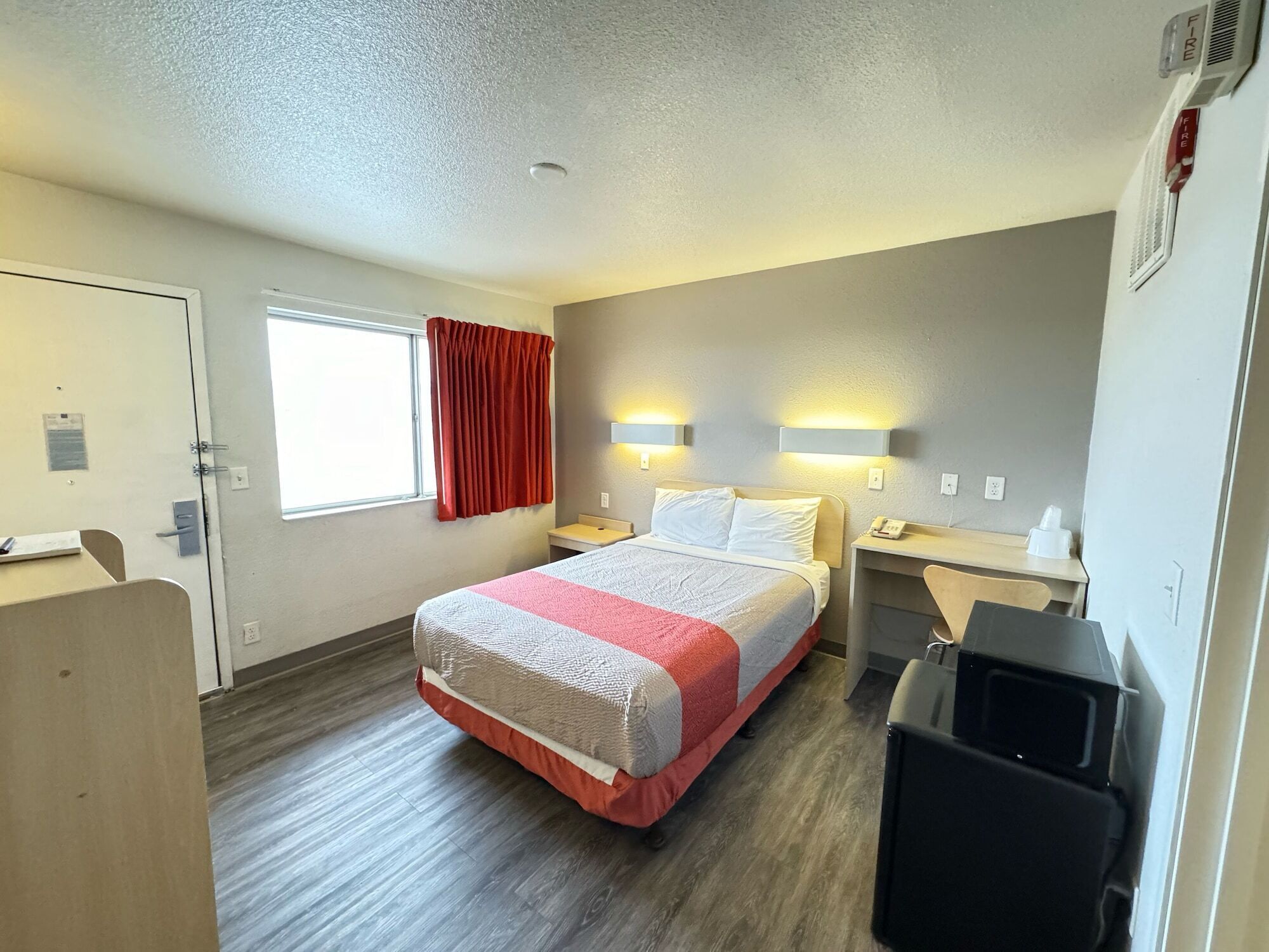A hotel room with a bed, bedside tables, a desk, a chair, a trash bin, a window with curtains, and a door.