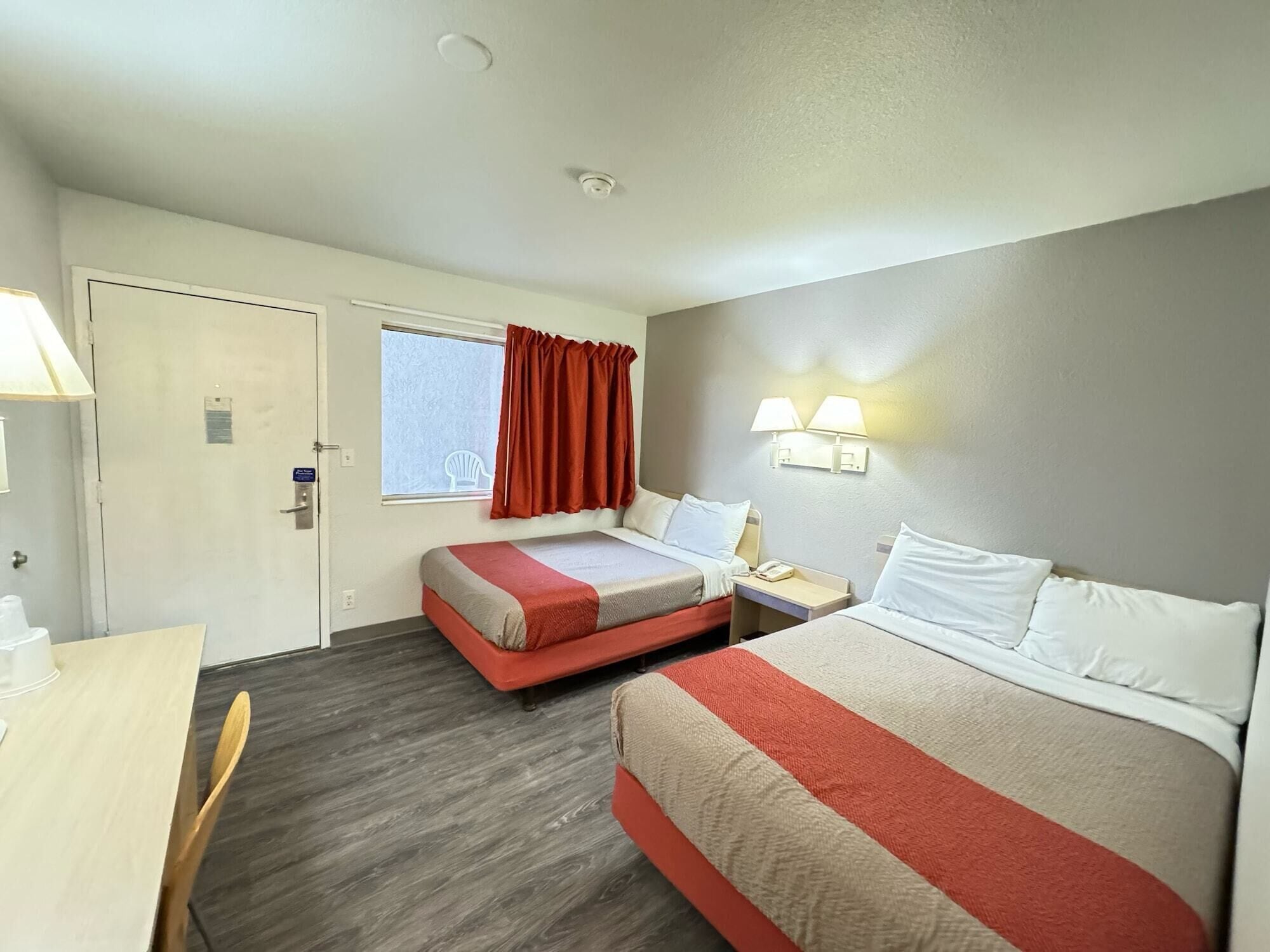 Une chambre d’hôtel avec deux lits, un bureau, une chaise, une fenêtre avec des rideaux et une lampe fixée au mur.