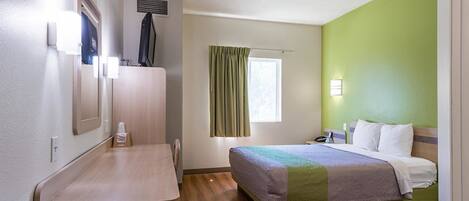 A hotel room with a bed, a desk, a chair, a window with curtains, a wall-mounted air vent, and a wall-mounted light.