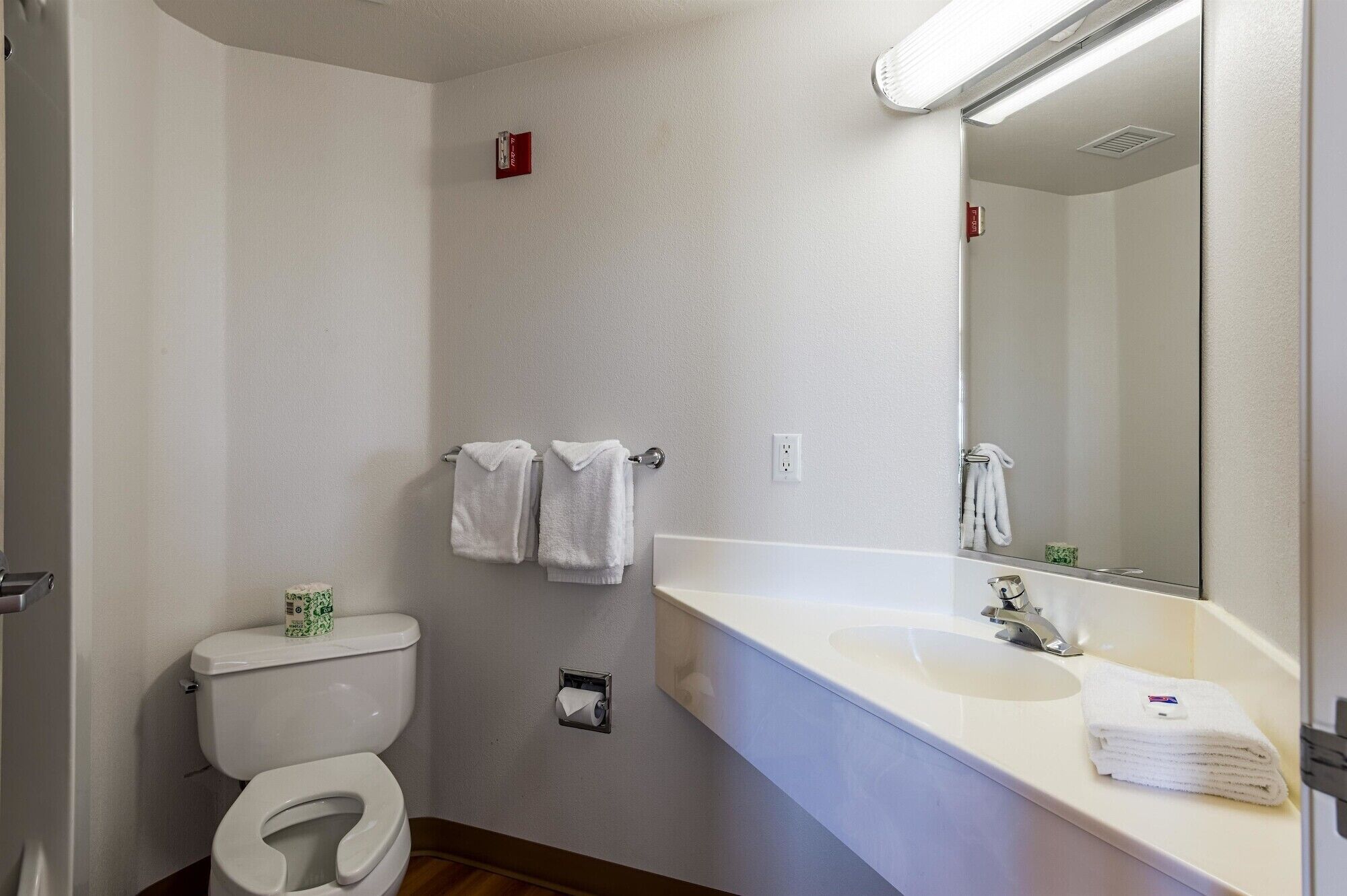 A bathroom with a toilet, sink, and mirror. There are towels hanging on the wall and a roll of toilet paper.