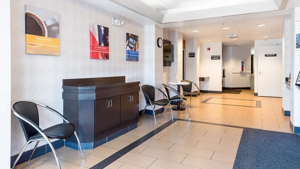 Un hall d’hôtel avec un comptoir de réception en bois foncé, des chaises noires et un sol carrelé. On peut voir des panneaux indiquant « Distributeur de glaçons » et « Ascenseur » sur le mur.