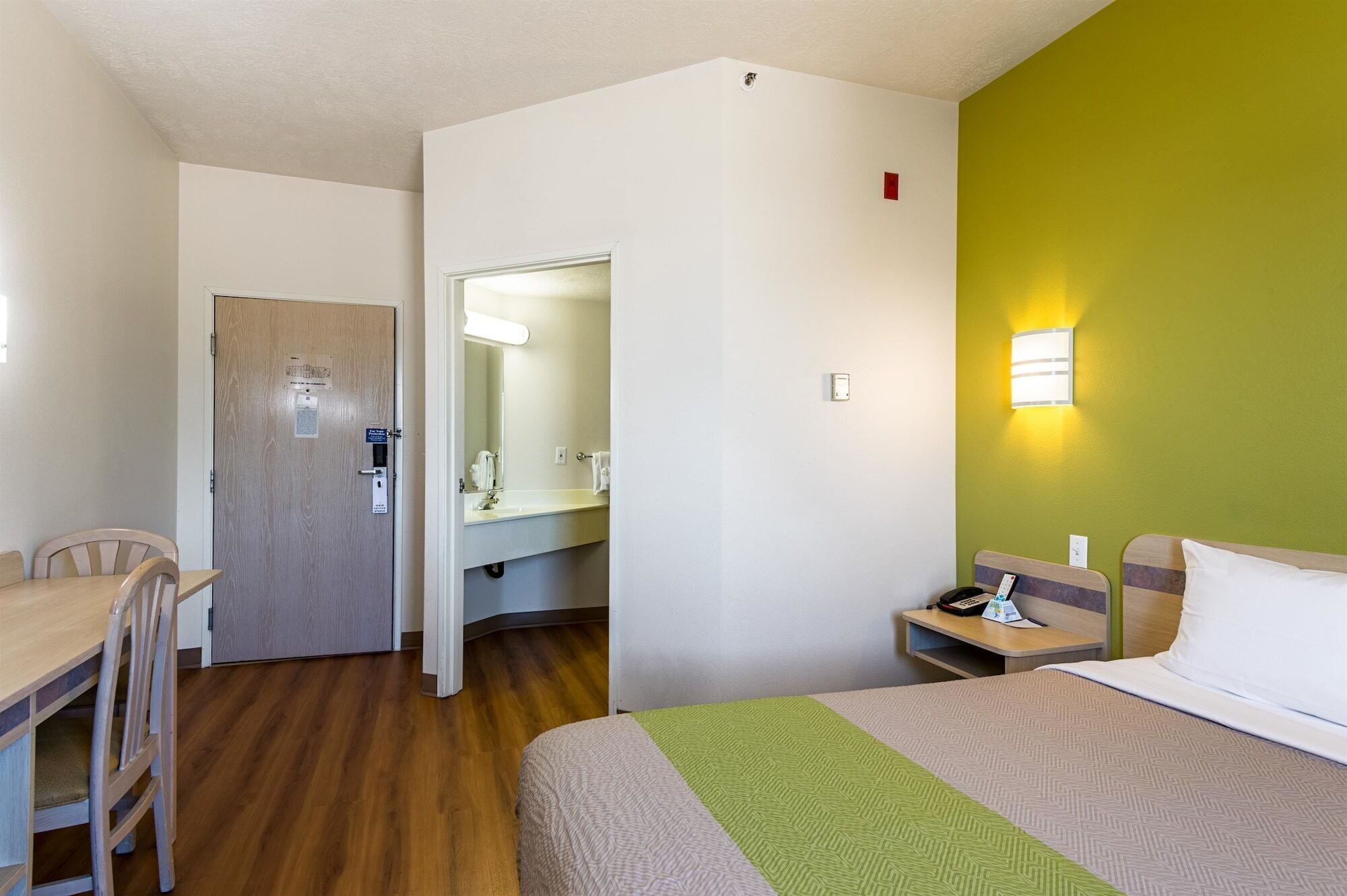 Habitación de hotel con cama, escritorio y silla, baño con espejo y pared verde.