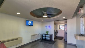 A reception area with a ceiling fan, a mounted television displaying 'America's Best Value Inn', a wall-mounted air conditioner, a reception desk with a plant, and a tiled floor. - Americas Best Value Inn Amarillo Airport (Amarillo)