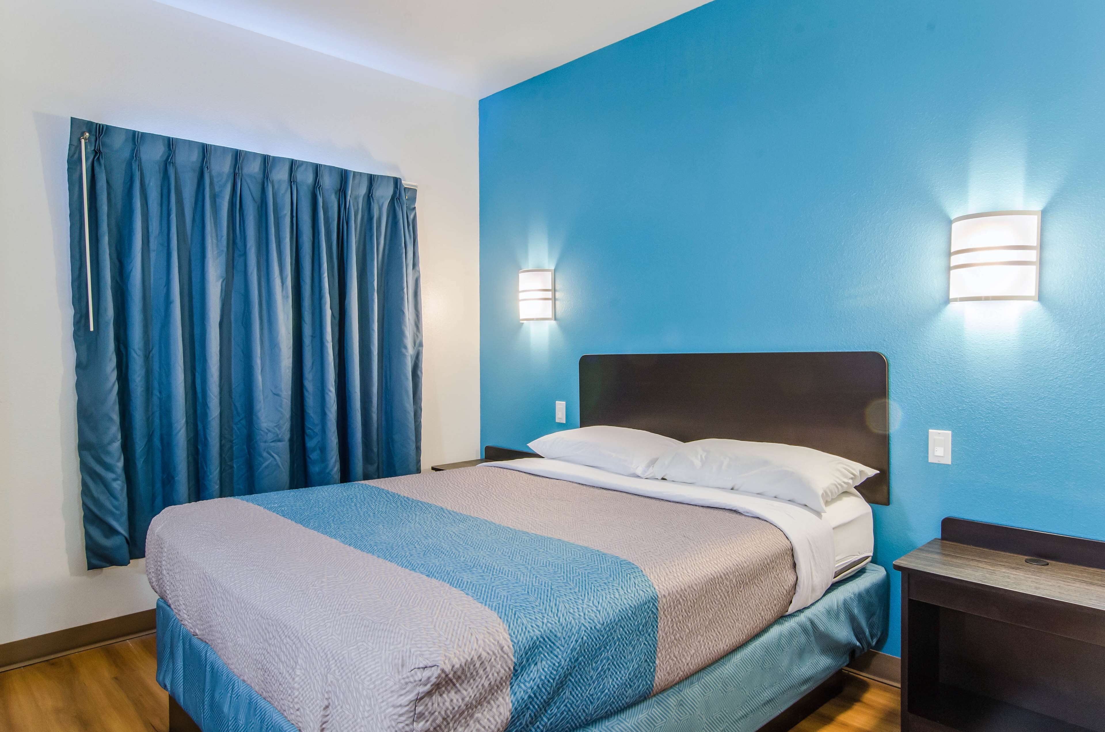 Una cama bien hecha con una colcha azul y blanca, dos luces fijadas a la pared y una cortina azul.