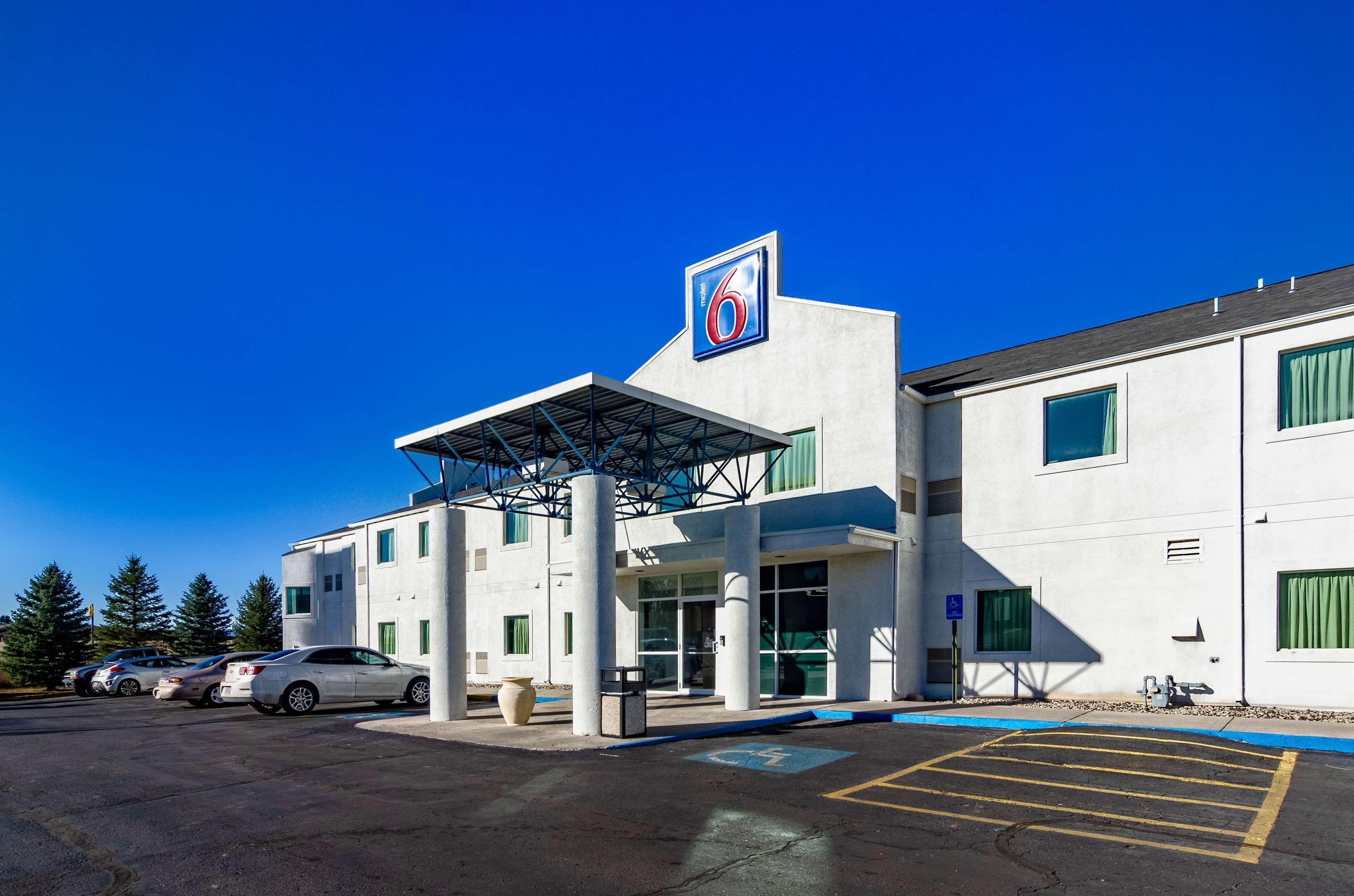 A modern building with a prominent '6' sign, a covered entrance, and a parking lot with several cars.