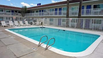 Une piscine dotée d’une terrasse blanche, entourée de chaises longues et bordée d’un bâtiment avec des balcons.