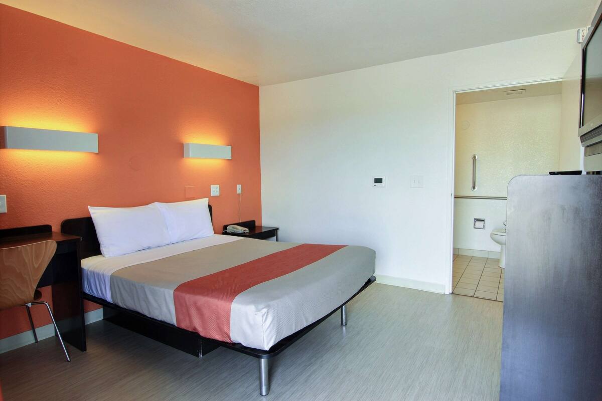 Une chambre d’hôtel moderne, équipée d’un lit, de tables de chevet, d’une chaise et d’une salle de bain visible à travers une porte ouverte.