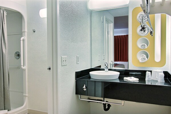 A bathroom with a sink, mirror, and shower area. - Motel 6 Corpus Christi, TX - East - North Padre Island (Corpus Christi)