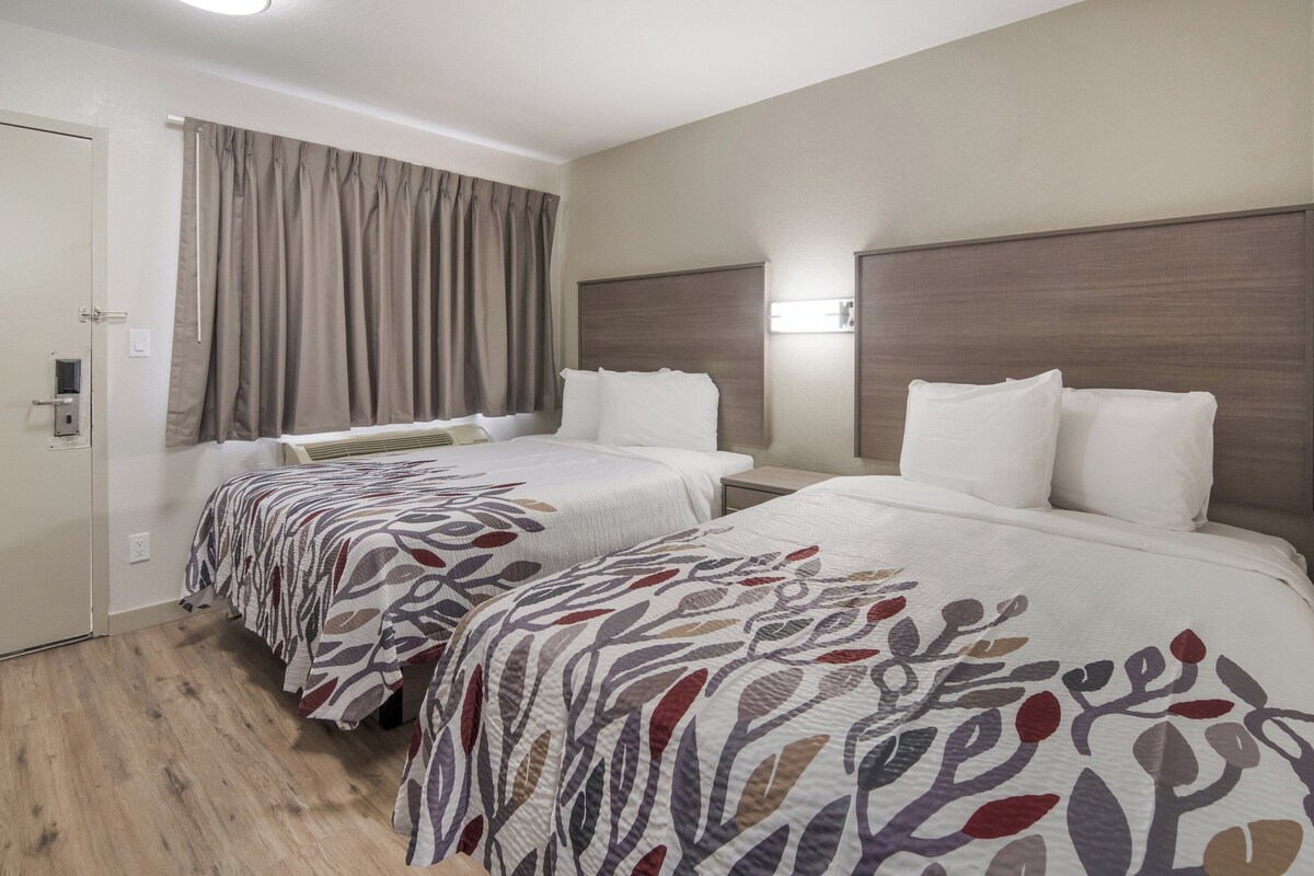 A hotel room with two beds, a large window with curtains, a wooden floor, and a wall-mounted light fixture.