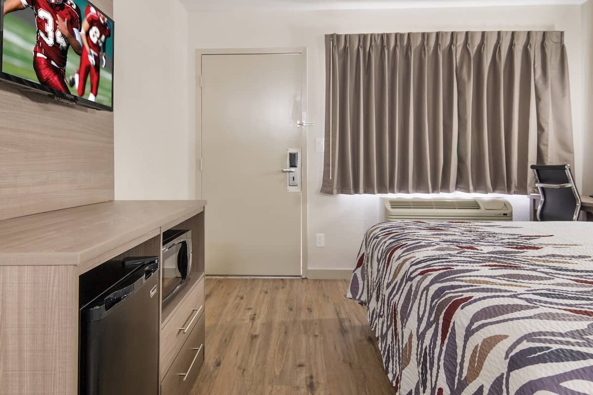 A hotel room with a bed, a TV mounted on the wall, a door, a window with curtains, and a small desk with a chair.