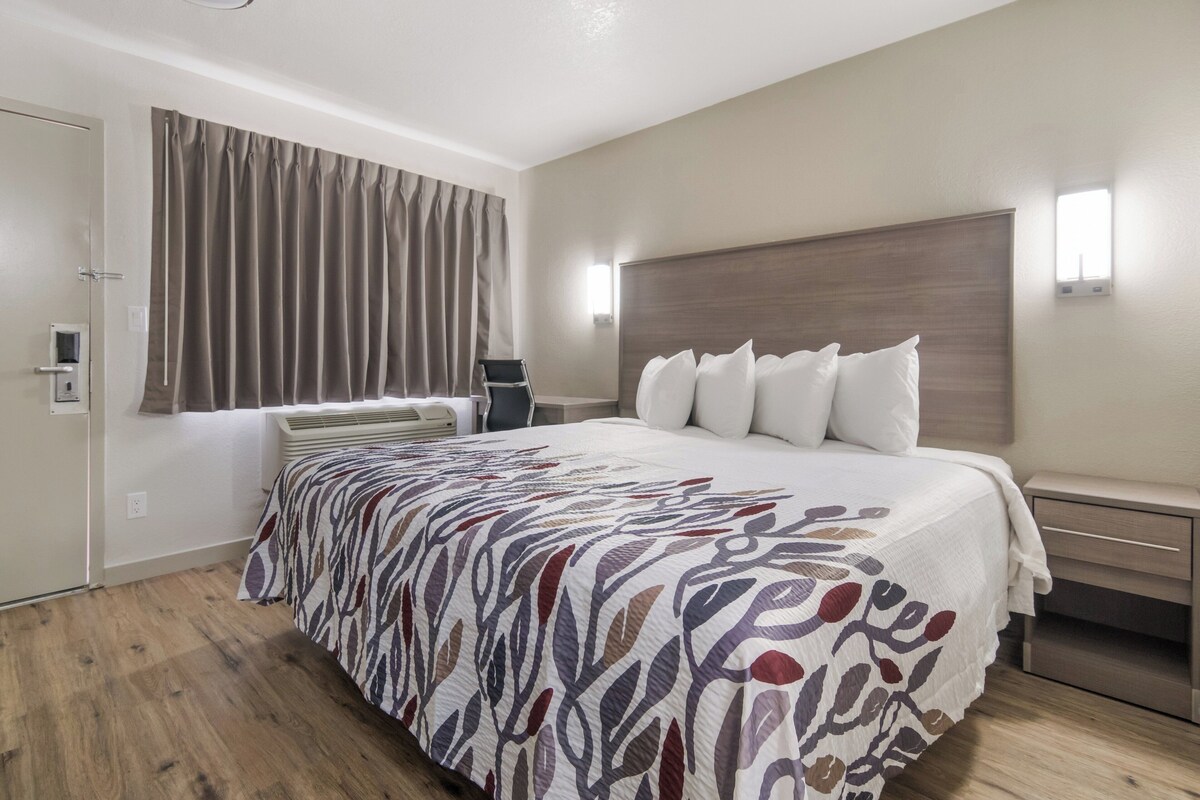 A hotel room with a large bed, a bedside table, a chair, a window with curtains, and a wall-mounted light.