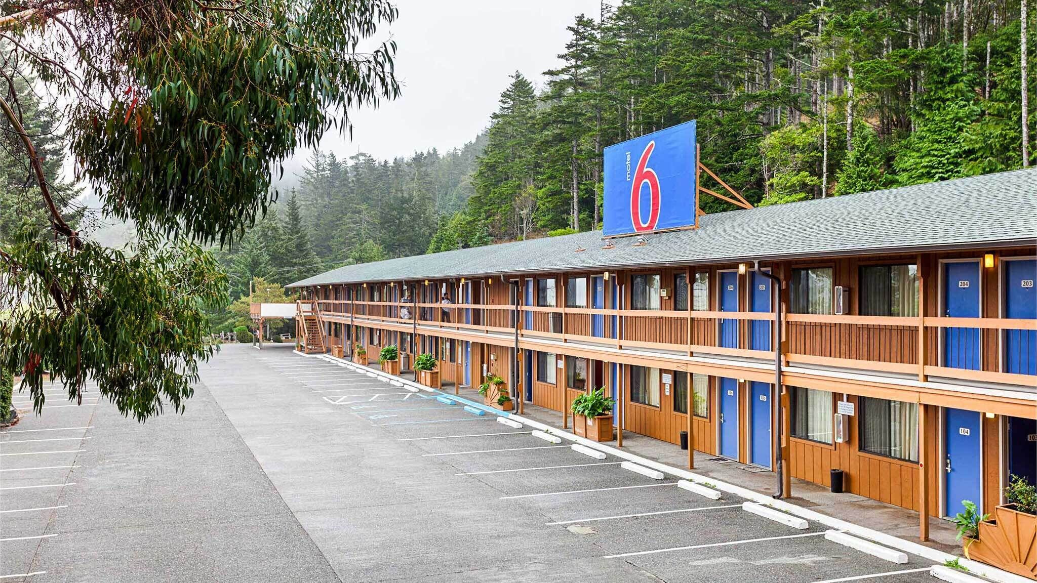 A motel with a row of rooms, a covered parking area, and a forested backdrop.