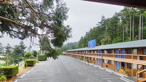 A motel with a row of rooms, a parking lot, and a forested backdrop. - Motel 6 Gold Beach, OR (Gold Beach)