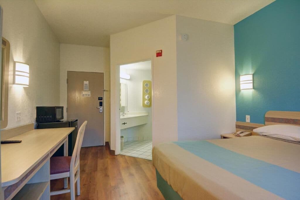 A hotel room with a bed, a desk, and a chair. There is a bathroom visible through an open door.
