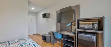Un dormitorio moderno con una cama, un escritorio, una silla, un televisor, un microondas y un refrigerador pequeño.