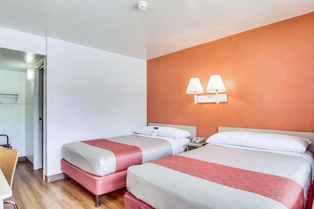 A hotel room with two beds, a wooden floor, a white wall, and a red accent wall.
