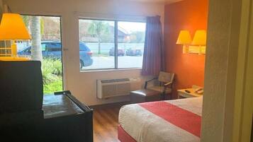 Habitación de hotel con cama, dos lámparas, una silla, un escritorio, una ventana con vistas a coches aparcados y un calentador.