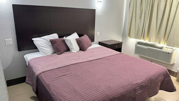 A bed with a headboard, pillows, a bedside table, and an air conditioning unit. - Xpress Inn & Extended Stays (Marshall)