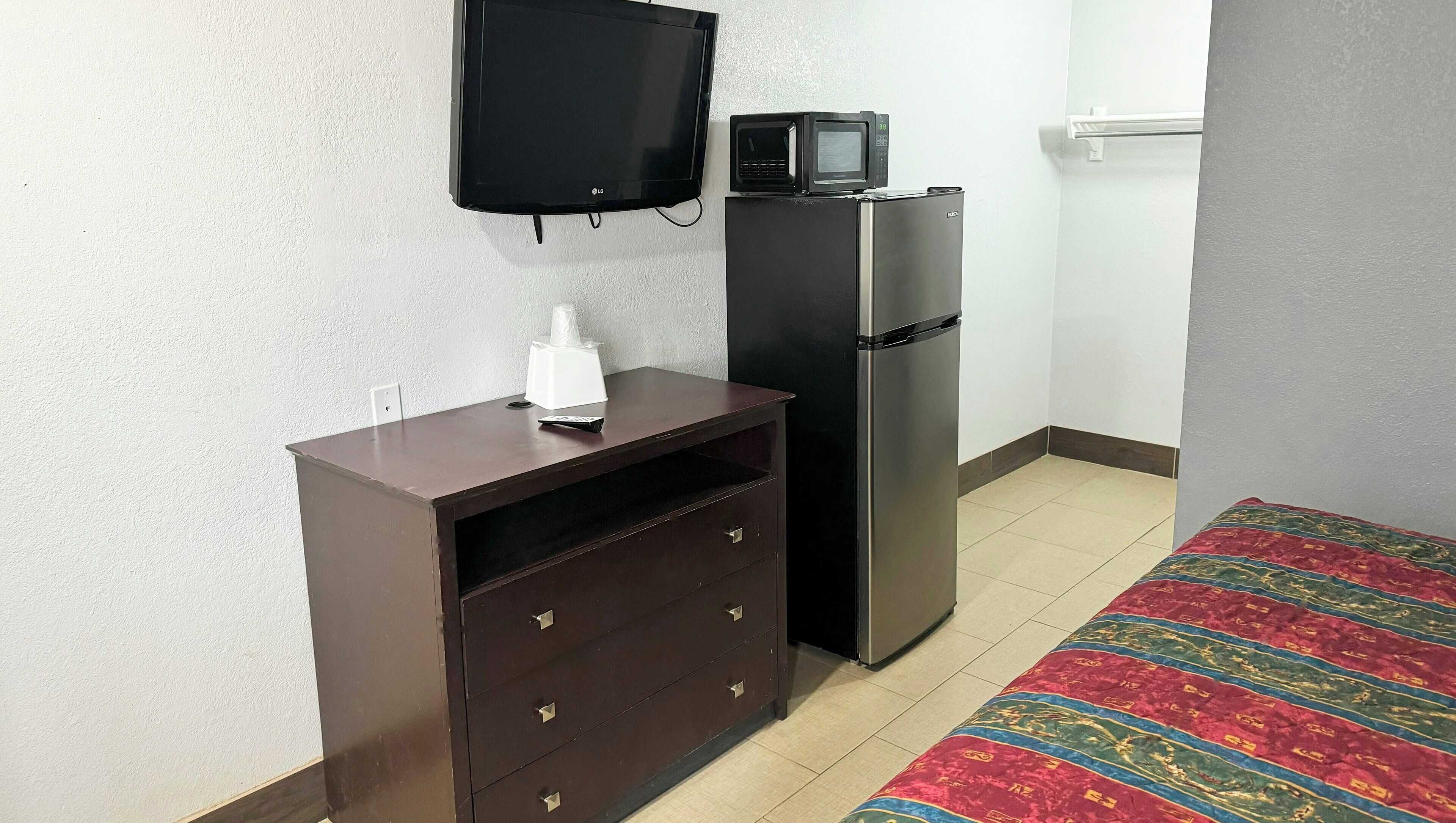 A room with a TV mounted on the wall, a refrigerator, a wooden cabinet with drawers, and a patterned rug on the floor.