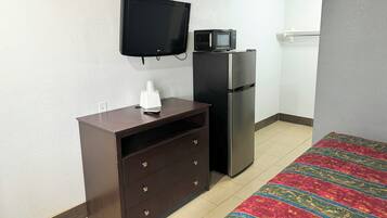 A room with a TV mounted on the wall, a refrigerator, a wooden cabinet with drawers, and a patterned rug on the floor.