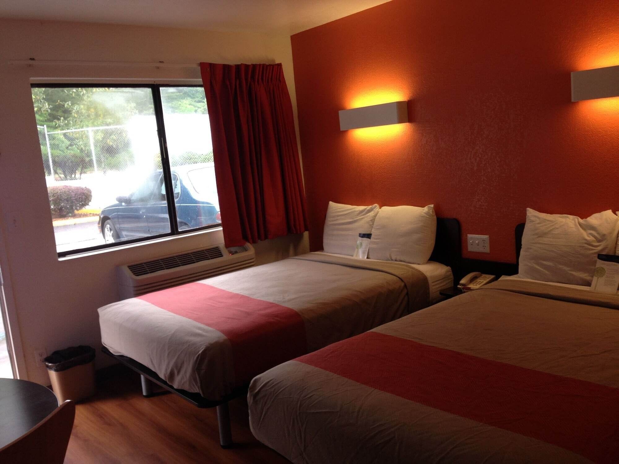 A hotel room with two beds, a window with a view of a parked car, a wall-mounted light, and a heater.