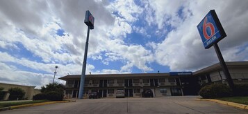 Un motel con un letrero que dice '6 ENTER', un letrero de 'Bienvenido' y un estacionamiento con coches.