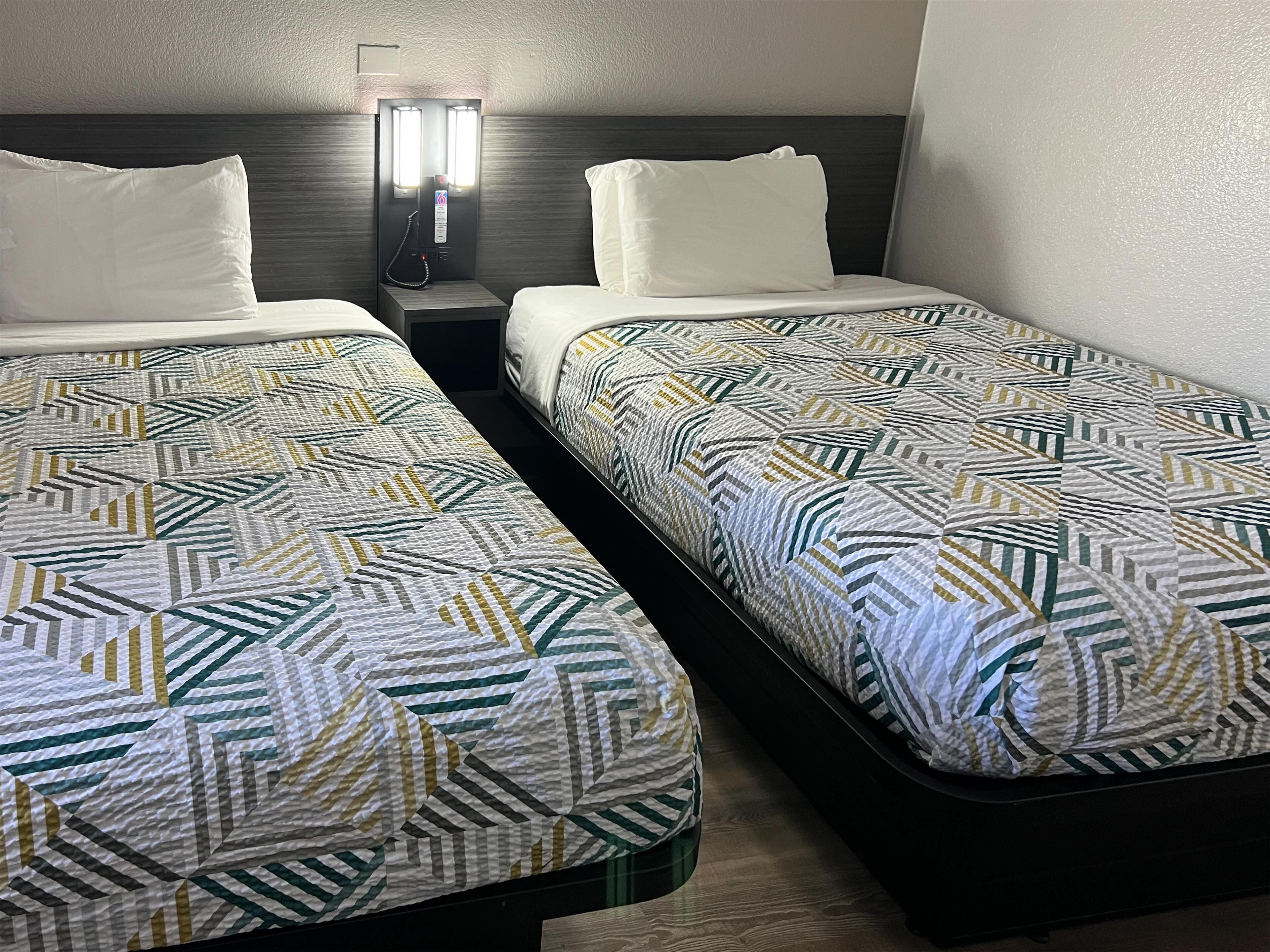 Two single beds with geometric patterned bedding, a bedside table with a phone, and a wall-mounted light fixture.