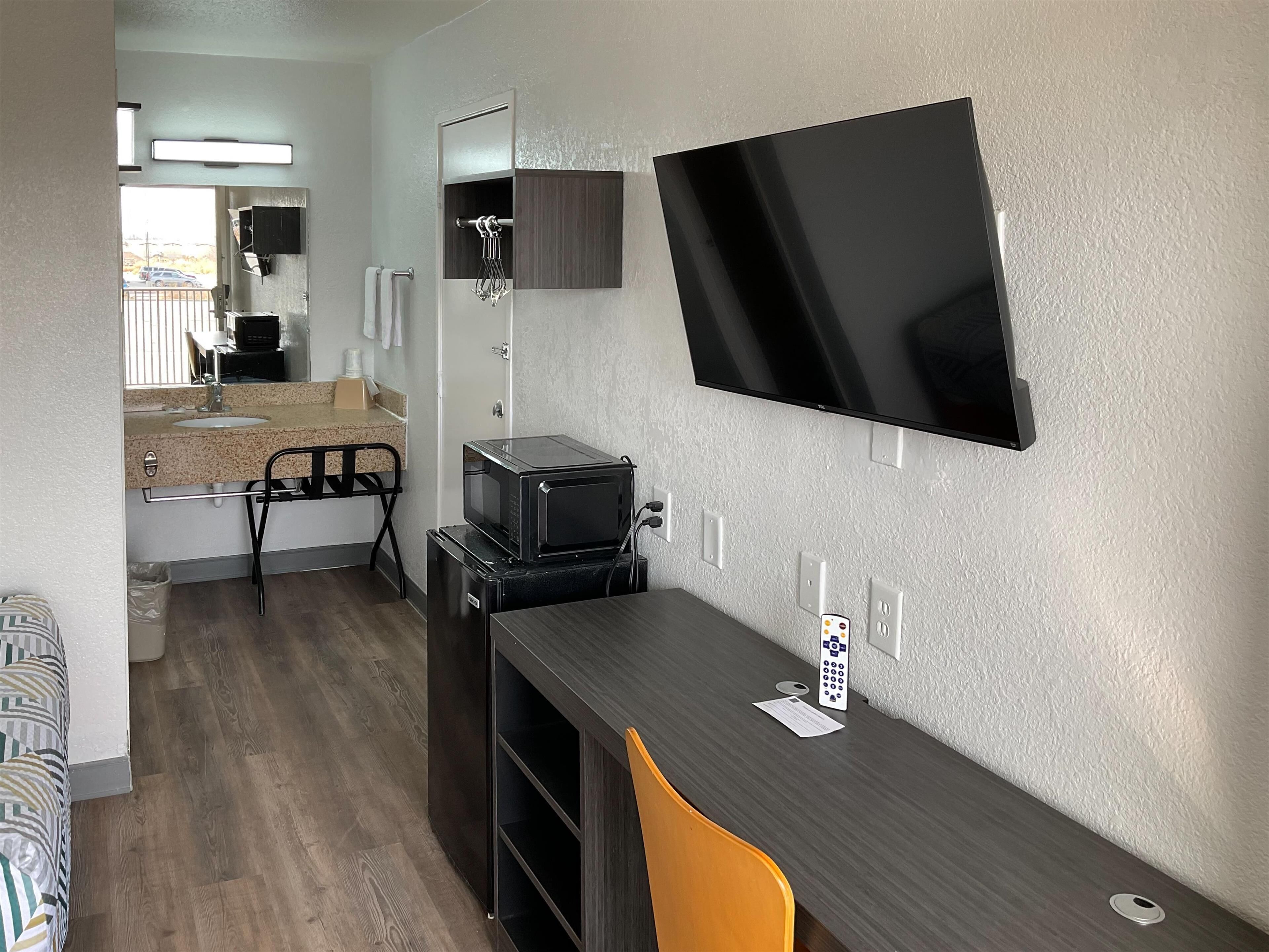 A compact hotel room with a kitchenette, a flat-screen TV mounted on the wall, a small desk with a chair, and a window offering a view of the outside.