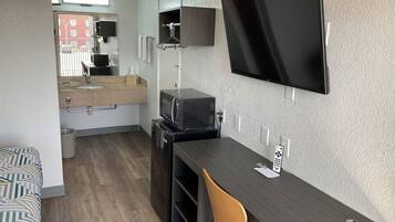 Una kitchenette compacta con microondas, lavamanos y perchero. Un televisor de pantalla plana está montado en la pared, y una silla de madera se encuentra ubicada en el escritorio.